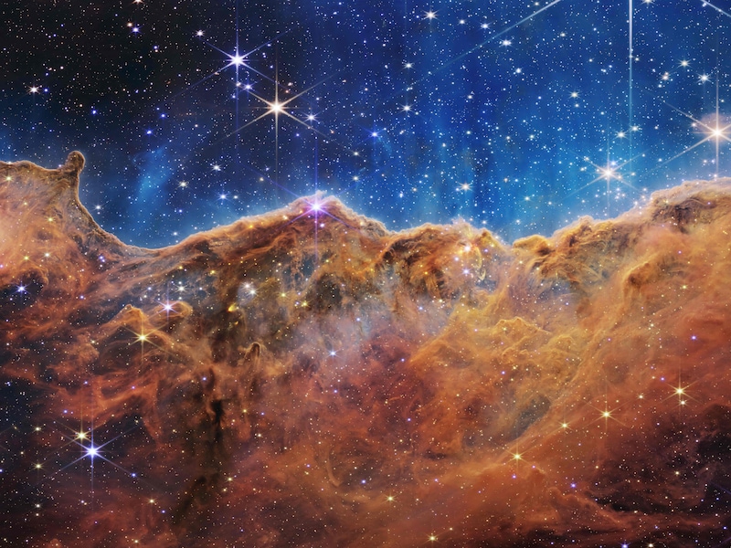 Een landschap van "bergen" en "valleien”, gespikkeld met glinsterende sterren dat eigenlijk de rand is van een nabijgelegen, jong, stervormingsgebied genaamd NGC 3324 in de Carina Nevel.