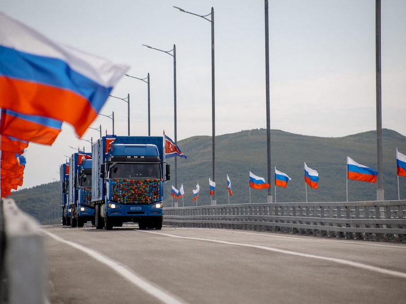Een arsenaal Russische vlaggen wapperend in de wind, terwijl een Noord-Koreaans konvooi vrachtwagens over de nieuwe brug komt gereden.