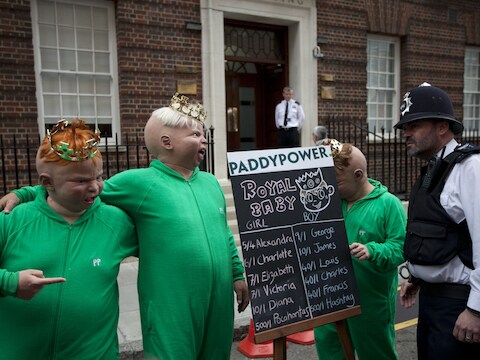 Medewerkers van een gokkantoor poseren met babymaskers voor een bord waarop potentiële namen voor de baby van Kate en William staan.