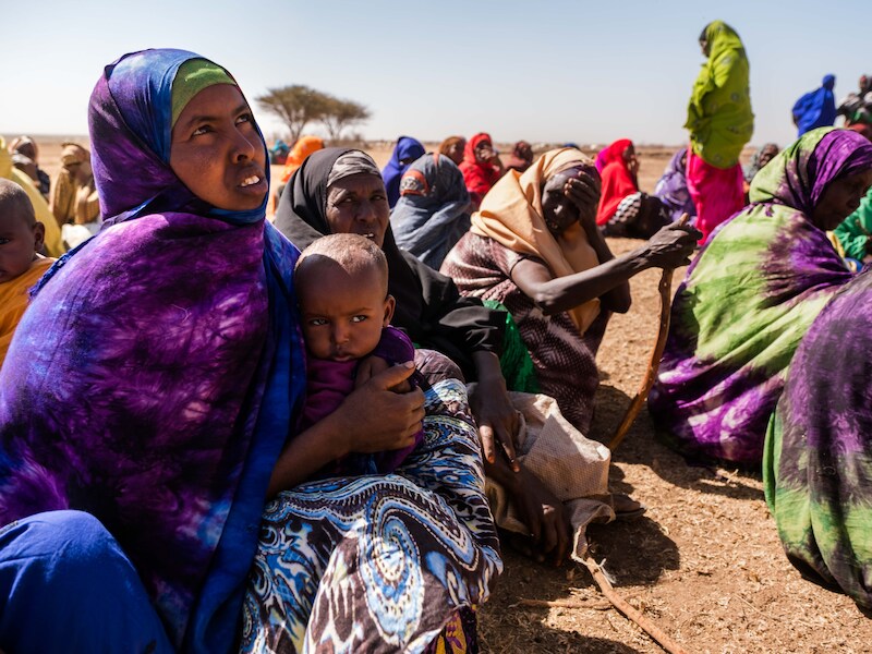 Vrouwen en kinderen wachten al zitten geduldig op de komst van de vrachtwagen. Op de voorgrond: Mama Sodoh met dochtertje Kadan.