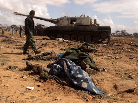 Een lid van de rebellen wandelt in de buurt van de stad Benghazi langs een gebombardeerde tank van het Libische regime. De lijken van gesneuvelde Kadhafi-getrouwe troepen zijn onder dekens gelegd.