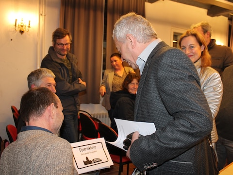 Sinds dat het bestuur eind januari 2026 bekendmaakte dat het twee gemeentezalen wil verkopen, zitten er quasi elke gemeenteraad inwoners uit de actiecomités van Hoogstade en Gijverinkhove in de zaal. Met een bordje ‘Gijverinkhove, dorp zonder dorps(t)huis’ als stil protest. Beeld: burgemeester Liefooghe passeert de actievoerders op het einde van de gemeenteraad.  