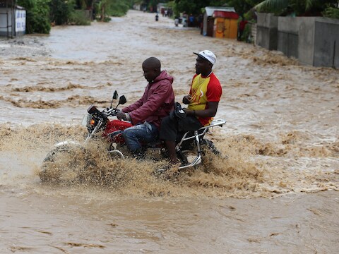 Mannen op een motorfiets steken een overstroomde straat over in Grand Goave.