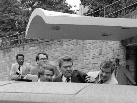 President Ronald Reagan zwaait en kijkt dan omhoog voordat hij door geheim agenten van de geheime dienst in de limousine van de president wordt geduwd na te zijn neergeschoten buiten een hotel in Washington, 30 maart 1981.