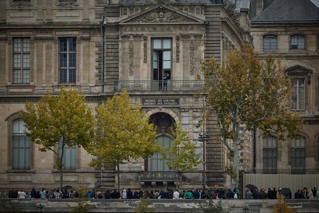 Ee menigte verzamelt zich voor de gevel waar de dieven hebben ingebroken in het Louvre, terwijl een agent foto’s neemt van de braaksporen op het raam.
