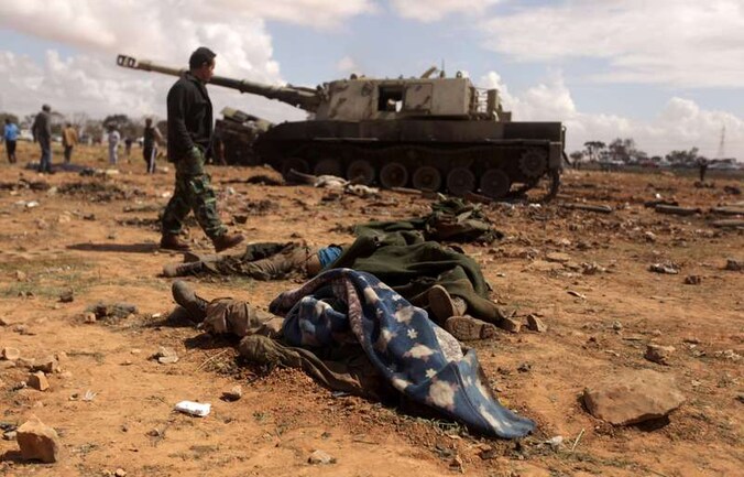 Een lid van de rebellen wandelt in de buurt van de stad Benghazi langs een gebombardeerde tank van het Libische regime. De lijken van gesneuvelde Kadhafi-getrouwe troepen zijn onder dekens gelegd.