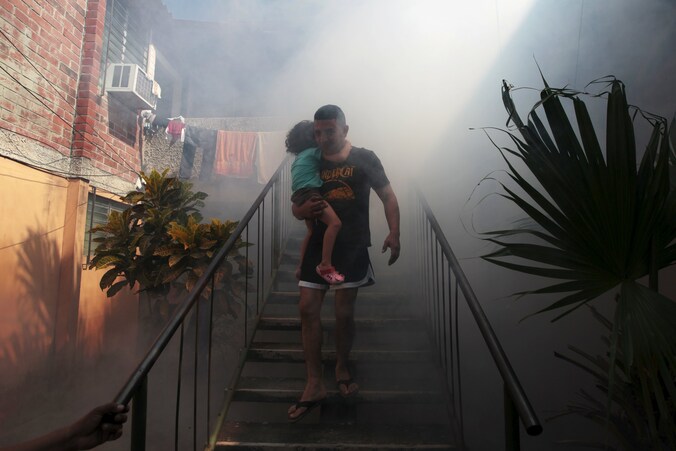 Een man draagt zijn zoontje op de arm terwijl gezondheidswerkers in Soyapango, El Salvador, verdelgingsmiddel spuiten tegen muggen om een verspreiding van het zikavirus tegen te gaan.