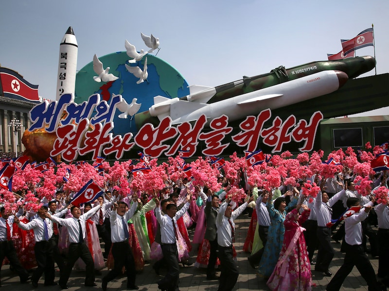 Noord-Koreanen zwaaien met de vlag van het land en plastic bloemen terwijl een vloot met raketten en het opschrift 'Voor vrede en stabiliteit in de wereld' voorbijkomt tijdens de militaire parade ter ere van de 'Dag van de Zon'.